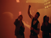 People dancing with lively expressions in a dimly lit, red-hued environment with a guitar graphic projected in the background.