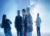 A group of four people interact energetically in front of a large screen displaying a guitarist.