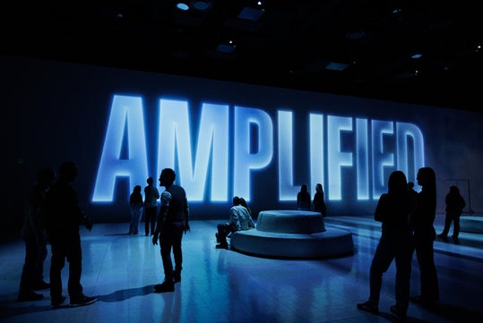 Large illuminated letters spell "AMPLIFIED" in a dimly lit room, with silhouettes of people standing and walking around.