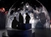 Three people sit and stand on circular platforms, silhouetted against a large, detailed image of the moon projected in the background.