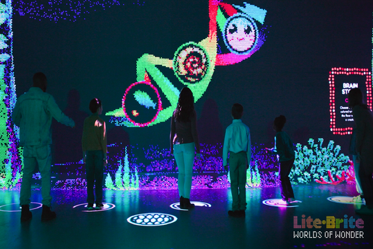 People interacting with a colorful digital art installation. Silhouettes stand on circular projections, facing a large screen displaying vibrant pixel art. Text: "Lite-Brite Worlds of Wonder.