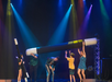 Performers on stage holding oversized magic wands under colorful spotlights, with the audience smiling and clapping.