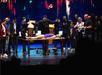 Magician performs illusion with woman on stage, surrounded by a group of onlookers. Stage lighting is colorful and dramatic.