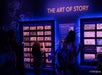 People observe and interact with illuminated storyboard displays under a sign reading "The Art of Story" in a dimly lit exhibit space.