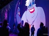 People stand in front of a large screen displaying an animated character with gray skin and red lips.