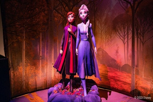 Two animated characters in detailed costumes stand on a rocky platform against a forest backdrop, with warm lighting and autumnal colors.