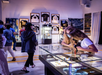 People wearing masks observe items displayed in glass cases inside a well-lit exhibition room with framed shirts and artwork on the walls.