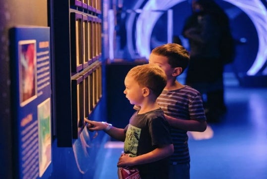 Two young boys interact with an illuminated display in a dimly lit, science museum-like setting.