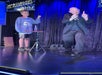 A magician in a suit gestures on stage beside a boy in casual clothes. They stand near a small table with a bottle; a sign above reads "IMPOSSIBILITIES" and stage curtains are visible behind them.