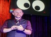 A man stands on stage holding papers, with a screen behind him displaying large cartoon eyes.