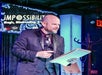 A man in a suit performs on stage with a notepad and pen, under a "Impossibilities" sign.