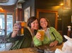 Two women sitting at a wooden table in a bar, smiling and holding up glasses of beer for a toast.