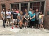 A group of thirteen adults pose for a photo together outside a brick building with windows and a door.