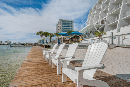 White Adirondack chairs facing the water on a wooden boardwalk, with a modern building, palm trees, and beach umbrellas in the background under a partly cloudy sky.