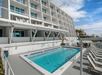Poolside view of a modern, white multi-story hotel with loungers arranged along the pool deck. Clear blue sky and distant buildings are visible in the background.