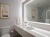 Modern bathroom with a large mirror, white countertop with a sink, toiletries, and towels. A framed picture hangs above the toilet. Glass shower enclosure in the background.