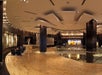 Spacious modern lobby with curved wooden floors, art displays, dark wall panels, and comfortable seating areas. Large ceiling lights illuminate the space.