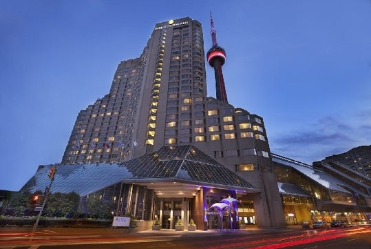 A tall hotel building with an adjoining tower is lit up at dusk. The street in front shows light trails from vehicles.
