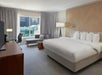 Hotel room with a large bed, TV, desk, and a seating area by a window with city views. Neutral tones and modern furnishings.