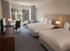 Hotel room with two double beds, a desk with a chair, a sofa, and a large window overlooking city buildings.