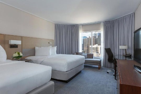 A hotel room with two double beds, a desk with a chair, a flat-screen TV, and a window showing a cityscape outside.
