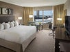 Hotel room with a large bed, pillows, a desk with a chair, a TV, a seating area, and a window with a city view.