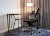 Modern office setup with a sleek black chair and chrome desk. A lamp and digital clock are on the desk. Gray curtains form the backdrop.