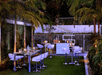 Outdoor patio setup with modern white tables and barstools on green grass, surrounded by palm trees and greenery, featuring a bar area in the background.