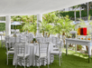Outdoor event space with round tables covered in white cloths, silver chairs, table settings, and a refreshment table with beverages, surrounded by greenery and palm trees.