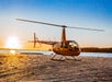 A small orange helicopter is parked on sandy ground near the water at sunset, with the sun low on the horizon.