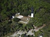 Aerial view of a black and white lighthouse surrounded by trees and a white fence, with a small building and cleared area nearby.