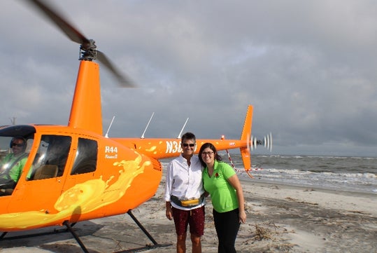 Two people stand on a beach in front of an orange helicopter, with another person visible inside the cockpit. The ocean and cloudy sky are in the background.
