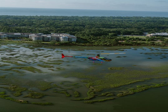 A colorful helicopter flies low over a marshland with apartment buildings and dense forest in the background near the coastline.