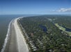 Aerial view of a long sandy beach bordered by dense green forest, residential homes, and golf course ponds under a clear blue sky.