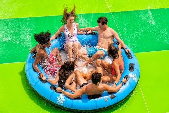 Five people sit in a blue inflatable raft, getting splashed as they ride down a bright green water slide.