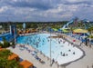 Aerial view of a crowded outdoor water park with a large wave pool, water slides, lounge chairs, cabanas, and people swimming and relaxing under partly cloudy skies.