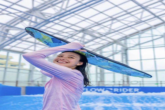 Person holding a surfboard overhead indoors, smiling, with a wave pool in the background.