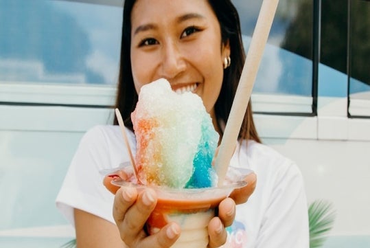 A girl is trying a colorful, cooling dessert of rainbow Shave Ice at Haleiwa as one of the Island and You stop.