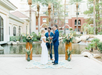 A couple stands at an outdoor altar with a person officiating their wedding. They are surrounded by floral arrangements and a water feature is in the background.
