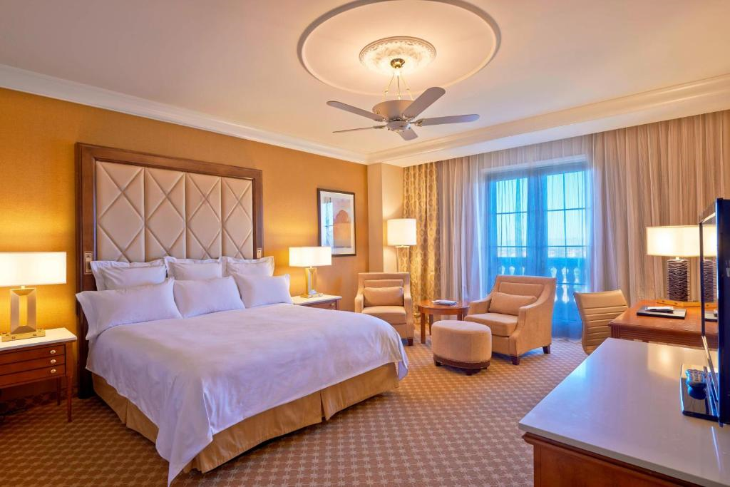 A well-lit hotel room with a king-size bed, two armchairs, a small table, and a TV. There is a ceiling fan, a large window with drapes, and warmly colored decor.