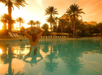 A person in a swimsuit stands in a pool at sunset, surrounded by palm trees, with a building in the background.