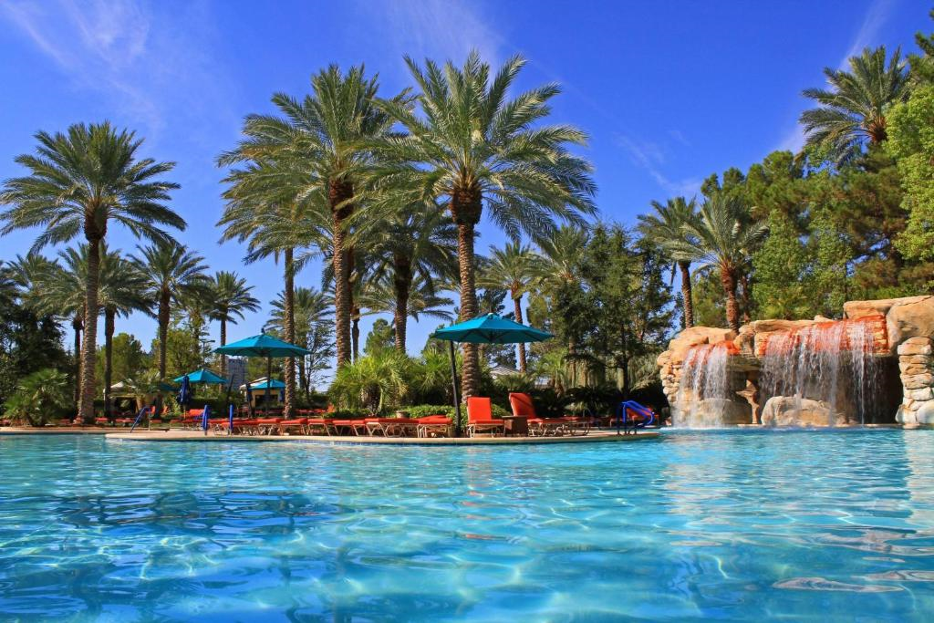 Outdoor swimming pool with palm trees, sun loungers, umbrellas, and a waterfall feature. Bright blue water reflects a clear sky. Relaxation area with a tropical ambiance.