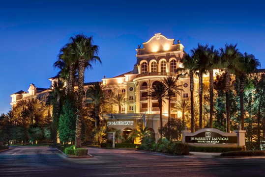 Hotel at night with illuminated facade, surrounded by palm trees. A sign reads "JW Marriott Las Vegas Resort & Spa" at the entrance. Clear evening sky in the background.