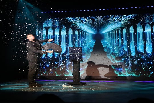 A magician performs on stage with a fan, scattering confetti, in front of a digital blue and black futuristic backdrop.