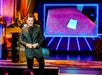 Jamie Allan smiling on stage with projection of glowing table illusion during Amaze Off-Broadway magic show in Midtown Manhattan.