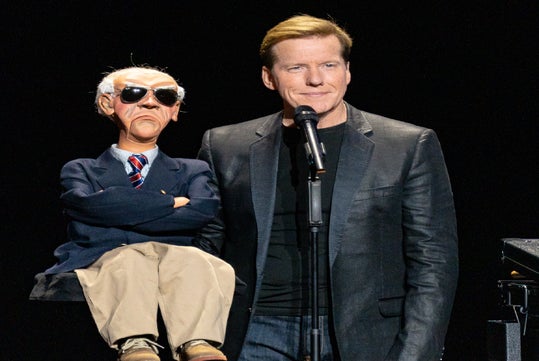 A man in a black jacket stands beside a ventriloquist dummy with sunglasses, a suit, and crossed arms, performing on stage with a microphone.