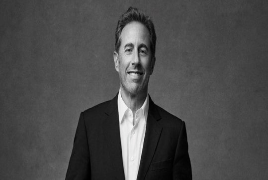 A man in a dark suit and white shirt stands against a plain gray background, smiling at the camera.