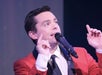 A man in a red jacket sings into a microphone on stage, raising both hands with fingers pointing upward.