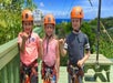 Three young kids in orange helmets and zip lining harness with their hangs doing the shaka sign on a sunny day.