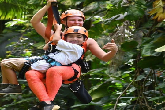 Two people wearing helmets and harnesses ride a zip line through a lush, green forest, smiling and enjoying the activity.